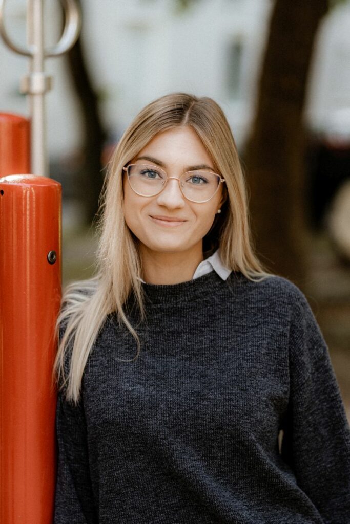 Junge Frau mit blonden Haaren und runden Brillengläsern, lächelt leicht in grauem Pullover, lehnt an rotem Metallpfosten.