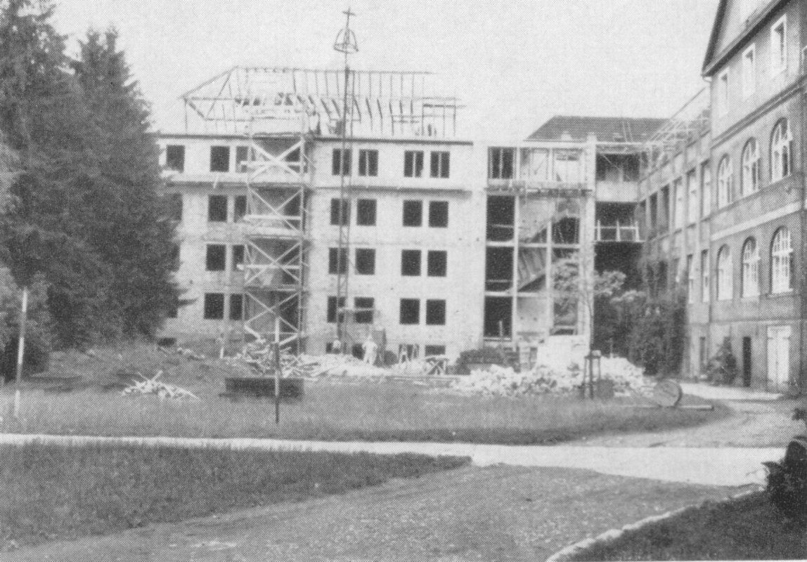 Mehrstöckiges Gebäude in Rohbau mit Gerüsten, Schutt und Baumaterialien auf einem Rasenplatz zwischen alten Backsteingebäuden.