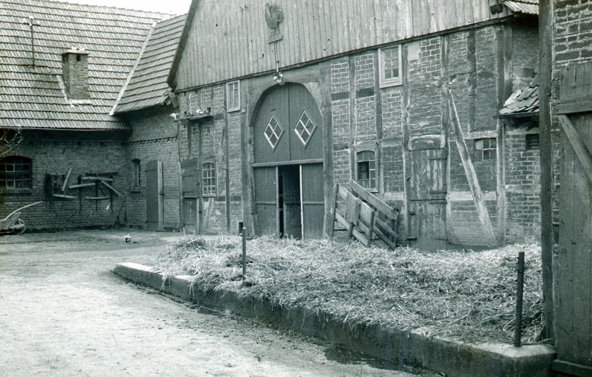 Innenhof eines alten Fachwerk-Bauernhofs mit großen Holztoren, Strohhaufen auf Betonboden und Werkzeug an der Wand.